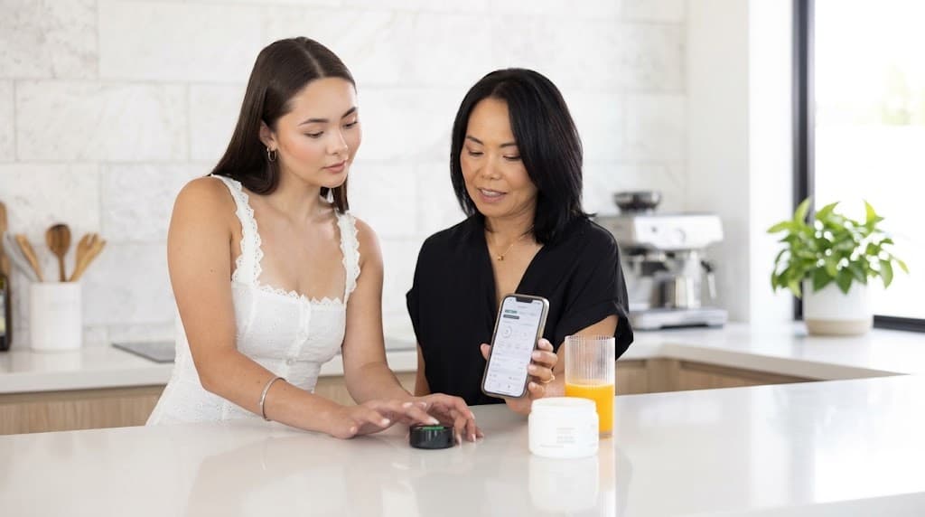 A pair of people at a kitchen worktop, one with a fingertip on the Prysm iO scanner and the other showing the phone screen
